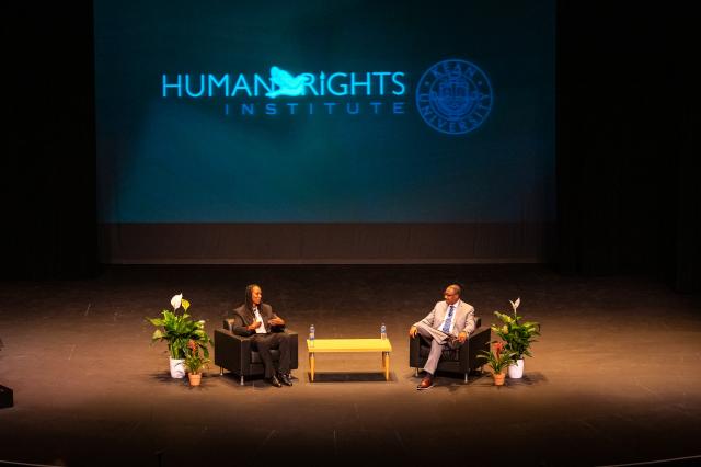 Two black people, a black woman and a black man, in business attire who are speaking to a crowd.