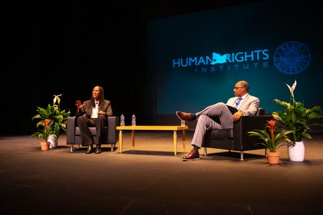 Two black people, a black woman and a black man, in business attire speaking to a crowd of people while sitting in chairs that are on a stage.