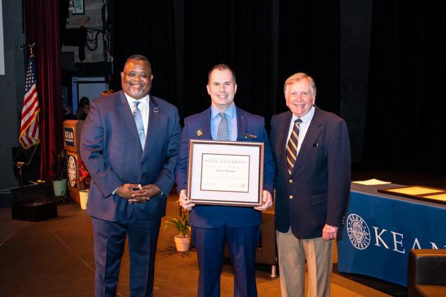 A group of men, a black man and two white men, in business attire are all standing next to each other and smiling for the camera. One white man is holding a plaque award.