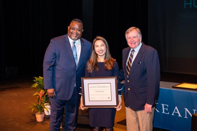 A group of people, a black man, an asian woman and a white man, all in business attire, are standing next to each other in front of a stage smiling for the camera. The asian woman is holding a plaque award.