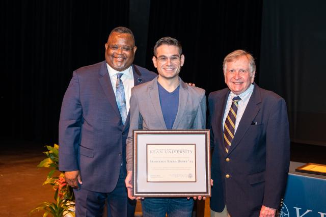 A group of men, one black man with glasses in a navy blue suit and two white men in suits, smiling at the camera in front of a stage. One white man in the middle is holding a plaque. 