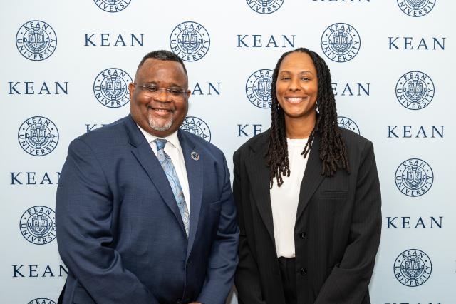 A black man with glasses and a black woman, both in suits, standing next to each other and smiling.