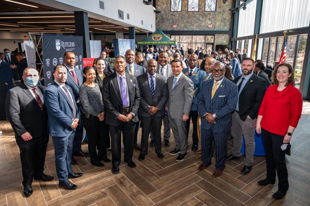 A group of people of all different ethnicities and races smiling and dressed in formal attire in a room with wooden floors and skylights.