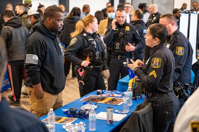 A black man with a black sweatshirt standing in front of a blue table listening to a white woman in police uniform speak to him.