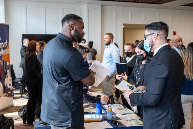 A black man in a black polo shirt speaking to a white man with glasses who is wearing a black suit.