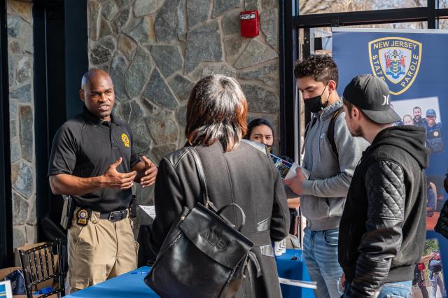 A black man wearing a black polo shirt with a yellow police force insignia on it speaks to a group of 3 people, one wearing a gray sweatshirt, the other a black backwards cap and black hoodie, and the last a woman holding a black purse and wearing a black peacoat.