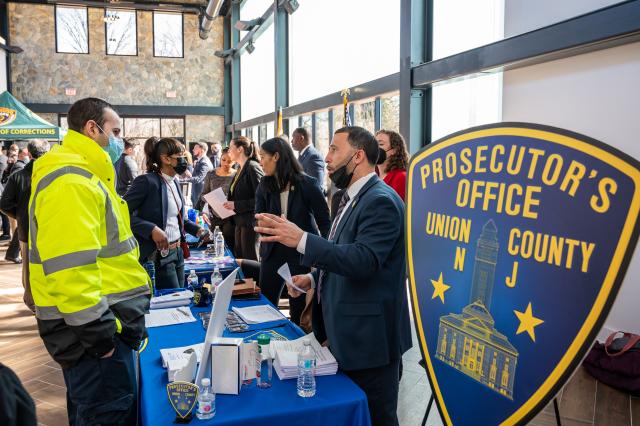 A tan man wearing a navy blue suit speaking to a white man in a fluorescent yellow coat. Both are standing next to a blue and yellow sign that reads: Prosecutor's Office Union County.