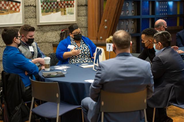 Six people of different ethnicities and races seated at a table with a Kean logo as the center piece, dressed in suits and talking to each other.