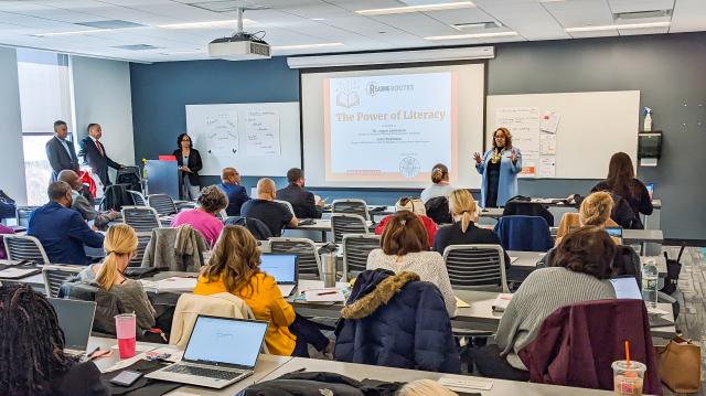 A brown woman with short, blonde hair in a light blue coat in a classroom full of students presenting about The Power of Literacy.