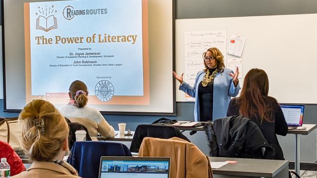 A brown woman with short, blonde hair and glasses wearing a light blue colored coat presenting a PowerPoint to a classroom full of students on The Power of Literacy.