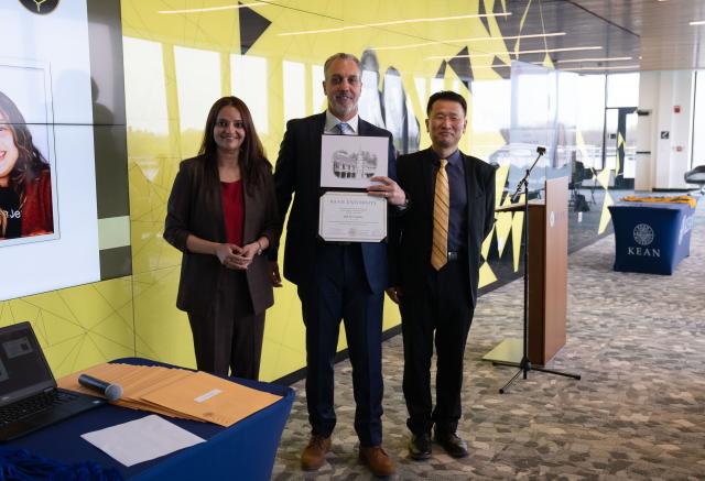 Two men and one woman, all wearing suits. The man in the middle is holding a certificate.