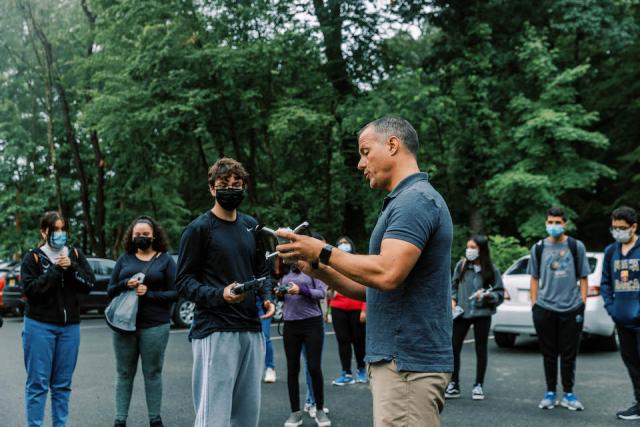 A man in a gray t shirt showing a young man in a black long sleeve shirt a remote control used to maneuver drones.