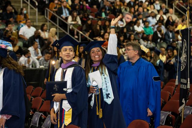 A woman in graduation cap and gown waves to someone in the audience.