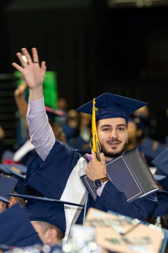 A male student in blue cap and gown stands and waves.