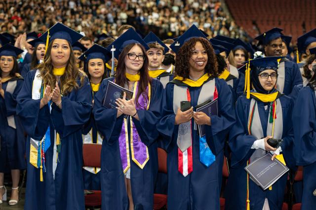 Four female students in blue caps and gowns, stand and applaud.