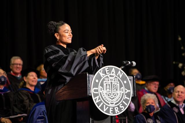 Actor Lauren Ridloff, wearing a graduation gown, clasps her hands, standing at a podium.