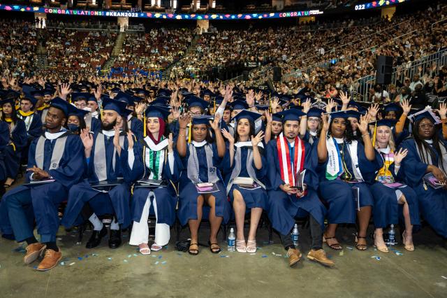 Students dressed in blue caps and gowns sit in a row, waving and smiling.