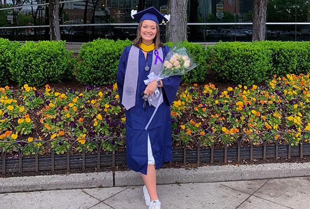 A young woman in blue graduation cap and gown, holds flowers as she stands in front of floral landscaping.