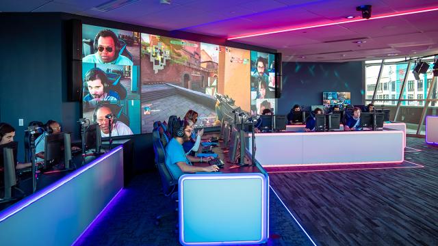 Students sit in rows before video gaming consoles, with a large screen display behind them.