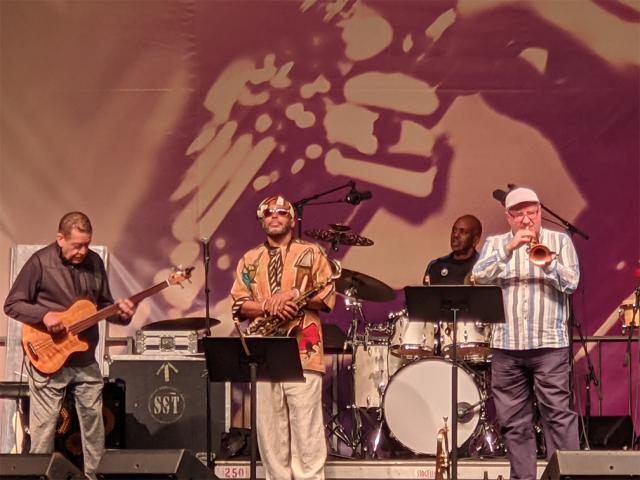 Four musicians on stage with the jazz and roots festival marketing image in the background