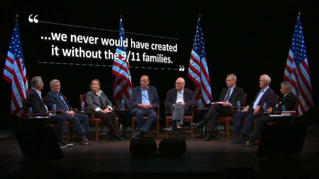 A group of people in a semicircle sit on stage. Overhead are the words, "we never would have created it without the 9/11 families."