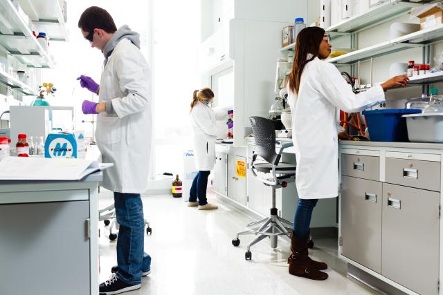 Three people with white lab coats and goggles work in lab.