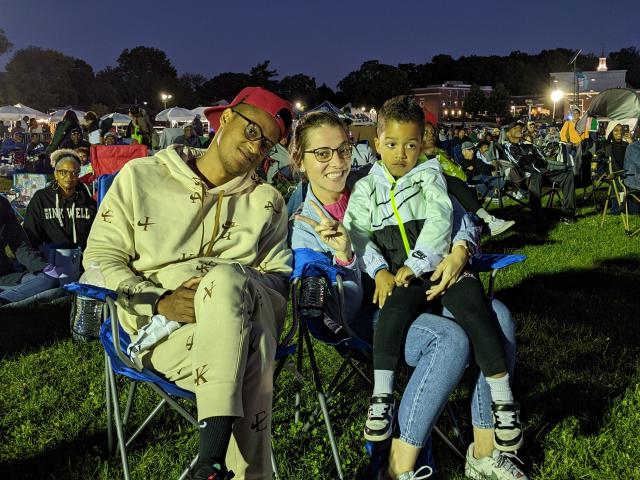 A young family sits on lawn chairs at the Jazz & Roots Festival. The mother gives a peace sign as her young son sits on her lap.