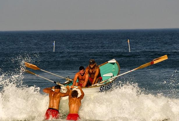 Four lifeguards, two in a rowboat and two pushing it, row out over ocean waves.