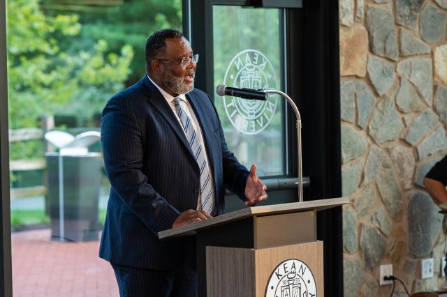 A photograph of President Lamont Repollet, a Black male wearing a dark navy suit and silver tie, is standing behind a podium with a microphone attached, speaking to members who attended this event. 