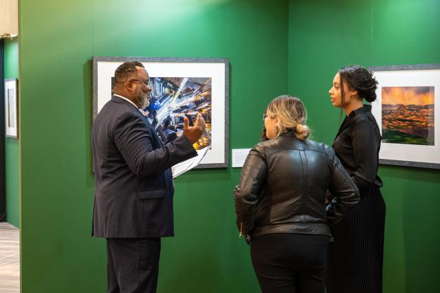 President Lamont Repollet, a Black male wearing a dark navy suit and silver tie, is standing with 2 females, who appear to be in their early 20’s, both wearing all black. The female closest to the President has white skin and dirty blonde hair in a bun, and the female to the right of her has darker skin,and dark black hair, also tied up in a bun. 