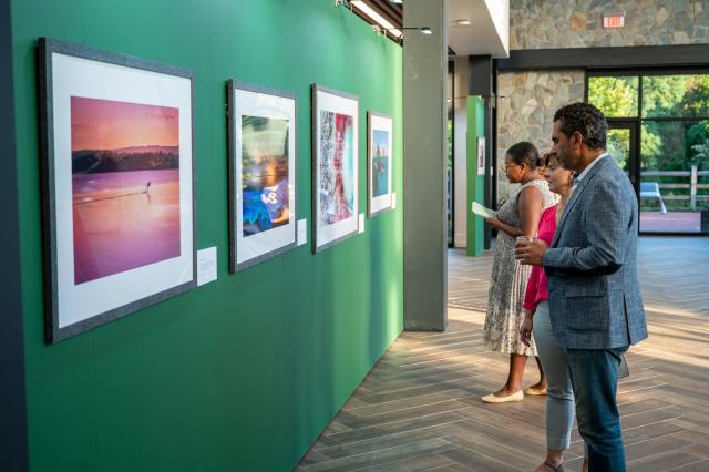 On the right of the picture stands a diverse group of men and women that work for Kean University, dressed in business professional attire, admiring the photographs that are on the left side of the picture, hung up on the wall to view.