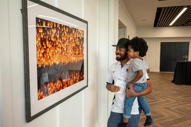 A Black male, with a black beard and mustache, wearing an all-black yankees baseball cap, and white collared shirt, is carrying a small Black male child, with black hair, gray t-shirt, jeans and black shoes. They are both admiring a photograph of orange lantern lights that are shining during the night-time.
