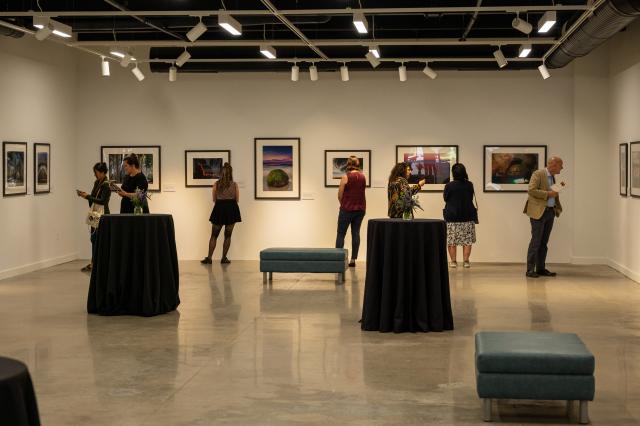 A wide shot of the photo gallery that displays framed photographs on the right and left walls, including the wall that is straight ahead. A diverse group of 6 females, and 1 male are admiring the photographs. Each individual is wearing business professional attire. The photo gallery has 2 tall, skinny tables with black table clothes touching the ground, that are spread out from each other towards the center of the room, and also holds 2 low-to-the-ground benches that are a lighter blue color.