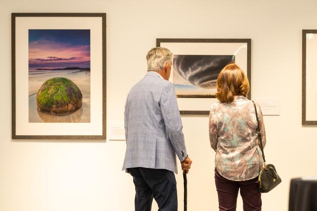 (L-R) a white male with gray hair, a light gray suit jacket, and dark pants, standing next to a white female with light brown hair wearing a paisley-designed top. Both have their backs to the camera admiring the photography on the wall. 