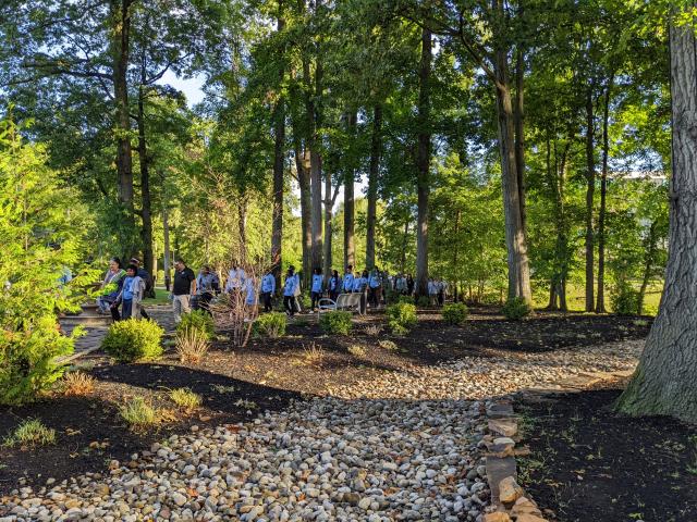 Wide shot of Kean students walking through tall trees. The sun is shining through, but the trees are providing shade. There is also a path full of small rocks that is parallel with the path the students are taking. 
