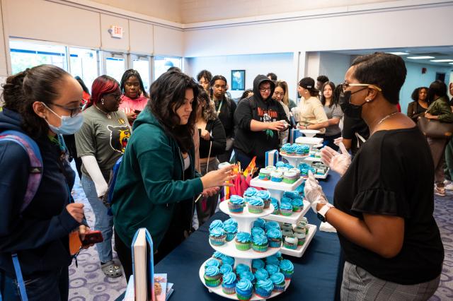 A wide shot of a Kean faculty member, a Black female with black hair, and glasses, wearing a black shirt and gray pants. She is behind tables that are holding cupcakes for students, and on the other side of the table are a diverse group of students waiting in line for a cupcake. 