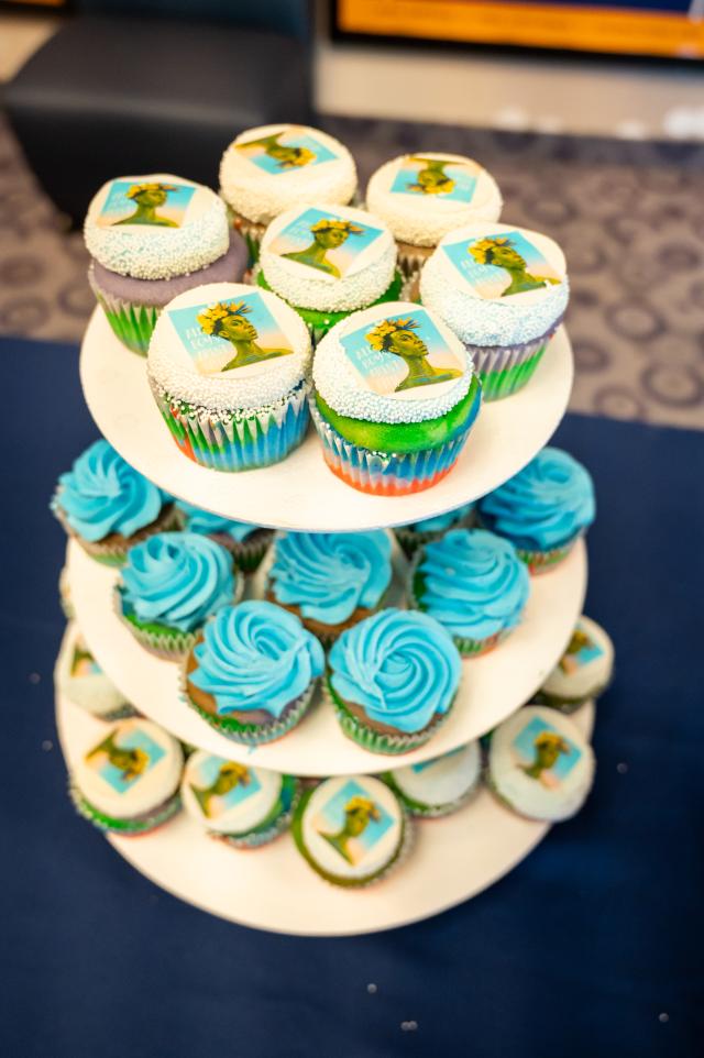 A 3 tiered turning table that is holding cupcakes. In the middle are rainbow cupcakes with blue icing, and on the top and bottom are rainbow cupcakes with vanilla frosting, and a picture of the book, All Boys Aren’t blue. 