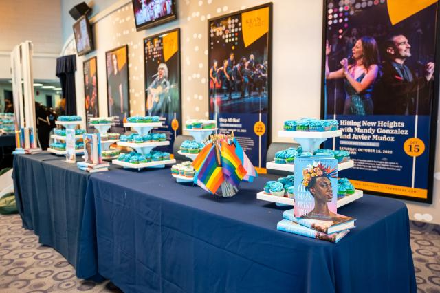  A wide shot of a 2 table long display that has 4 towers holding cupcakes, LGBTQ+ flags, and copies of the book, All Boys Aren’t Blue. The tables have navy table clothes on them.