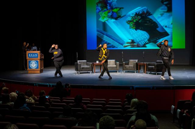 A wide shot of three Kean University students, Black males wearing black pants, black jackets, and black shoes, mid-dance as they perform for the crowd on stage. They are all lined up next to each other, spaced out. 