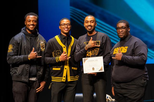 Author George M. Johnson is posing with three other Kean University students. (L-R) A Black male with curly short black hair, wearing black pants with a black jacket. A Black male with short curly black hair, wearing black glasses, black pants, and a black and yellow fraternity sweater. Author George M. Johnson, a Black male with black buzzed-cut hair wearing long black pants, a black crew neck with the year “1907” on it, and an uppercase “T” in the middle of the year in a mustard yellow color, holding a ce