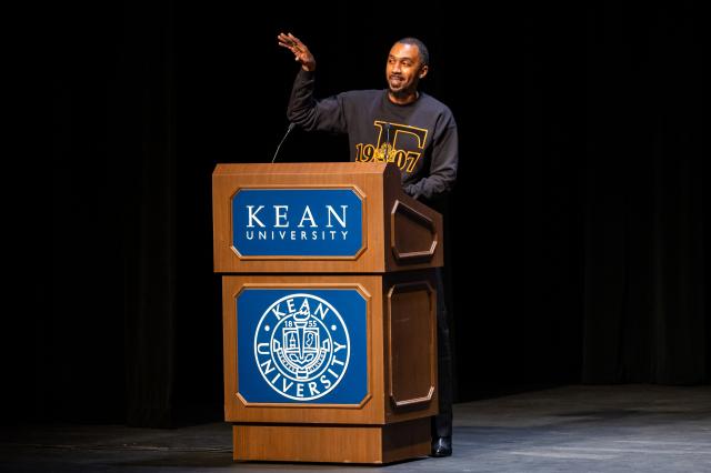 A close up shot of George M. Johnson,  a Black male with black buzzed-cut hair wearing long black pants, a black crew neck with the year “1907” on it, and an uppercase “T” in the middle of the year in a mustard yellow color, and black boots. They are standing behind a brown podium, with a Kean logo on the front, talking into a microphone, and gesturing to the crowd. 