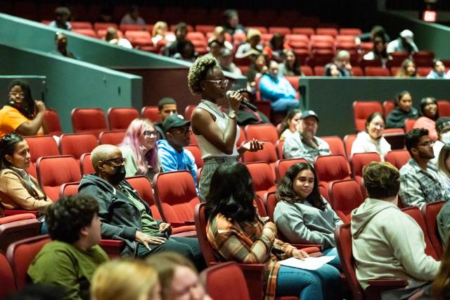 A wide shot of theater seats that are filled by a diverse group of students. One student, a Black female with curly black and blonde hair, wearing glasses and a white sleeveless turtleneck and plaid gray and tan pants, is standing in the middle of the crowd towards the stage, holding a microphone with her right hand, and speaking into it to ask the author a question. 