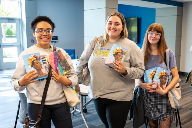 Photo of 3 Kean University students. On the left, a Black female with short black curly hair and black glasses, wearing a white crew neck, and black pants. In the middle stands a white female with short dirty blonde hair, wearing a gray long sleeve shirt and black leggings. On the right is a white female with long red hair and round, clear glasses, wearing a purple short sleeve shirt, and a black and white plaid skirt and black stockings. All 3 students are holding the book, All Boys Aren’t Blue, and smilin