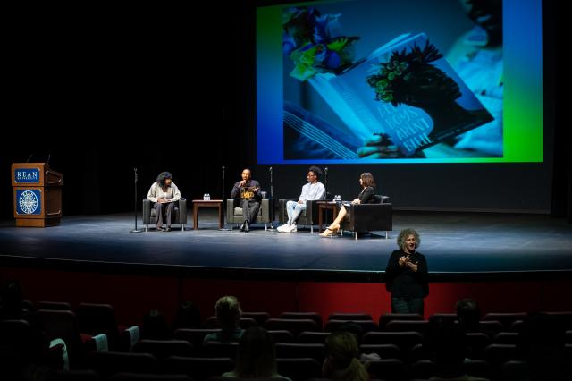 A wide shot of a stage that has 4 black chairs spaced out in a half circle, and each chair has a standing microphone next to it. From (L-R) seated is a Black female student with short black hair, wearing long gray, baggy pants, a light gray sweater with a white undershirt, and black slip ons, a brown chair with two small water bottles on the top of it is in between the next chair, who sits the Author, George M. Johnson, a Black male with black buzzed-cut hair wearing long black pants, a black crew neck with