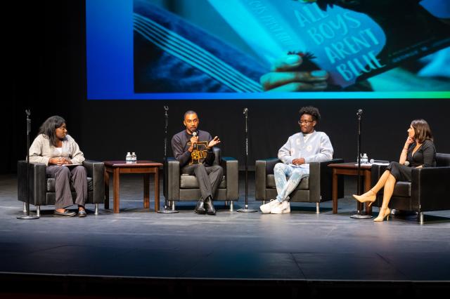 A wide shot of a stage that has 4 black chairs spaced out in a half circle, and each chair has a standing microphone next to it. From (L-R) seated is a Black female student with short black hair, wearing long gray, baggy pants, a light gray sweater with a white undershirt, and black slip ons, a brown chair with two small water bottles on the top of it is in between the next chair, who sits the Author, George M. Johnson, a Black male with black buzzed-cut hair wearing long black pants, a black crew neck with