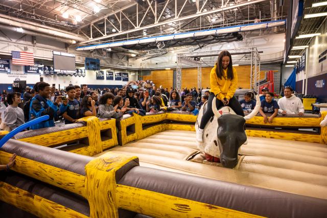 A Kean University student, a Black female with long brown hair in a yellow sweatshirt, black leggings, and white Nike air forces with white Nike socks showing, is on a blow-up mechanical bull. Students, alumni, and guests are surrounding the blow-up mechanical bull arena. They are holding up their phones and recording the student who is riding the bull. This is set up in the Kean Harwood arena, where the basketball games are held.