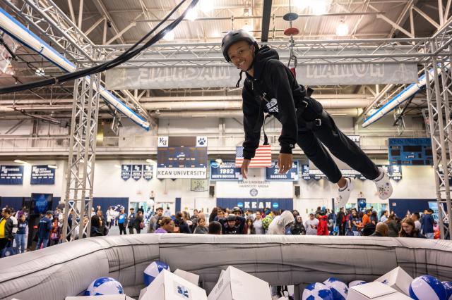 A Black female, wearing a black Kean University sweatshirt, and black sweatpants with white crocs, is harnessed and dangling above a huge ball pit, filled with white and blue Kean beach balls, and white mystery prize boxes. The female is wearing a black helmet, smiling at the camera. This is set up in the Kean Harwood arena, where the basketball games are held. Students, alumni, and guests are surrounding the ball pit, and the arena.