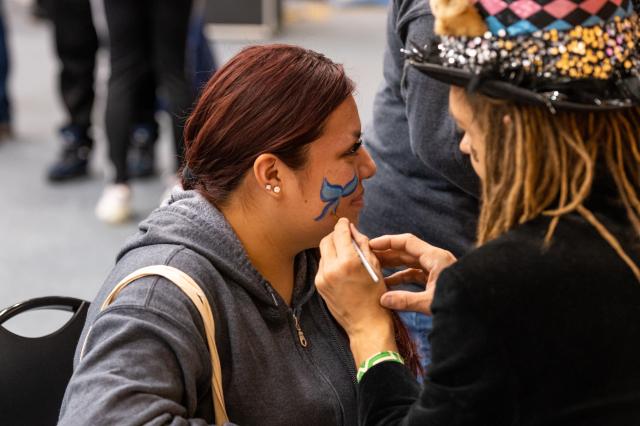  A Latinx female with red tinted hair tied back in a ponytail, is showing the right side of her face being painted. The face paint artist, a white male, or female (only their hand is shown), wearing a black jacket and tall black hat with feathers and sequins, seems to be drawing either a navy and baby blue butterfly or flower. The painting is not completed yet.