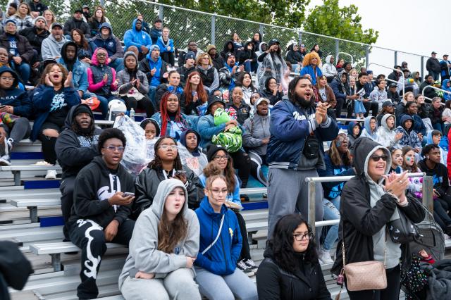 A wide shot of a diverse group of Kean students, alumni, parents, and fans that have filled the bleachers to watch the football game. They are wearing jackets and sweatshirts, and are cheering and clapping. 