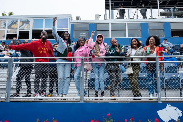 A diverse group of male and female Kean students wearing sweatshirts and jackets are lined up side-by-side leaning forward against the fence that lines the bleachers, smiling and cheering on the Kean Cougars football team.